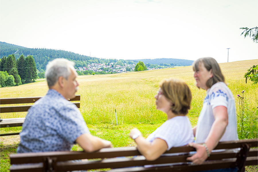 Rückansicht von drei Menschen, die auf einer Bank an einer Wiese sitzen und sich unterhalten.